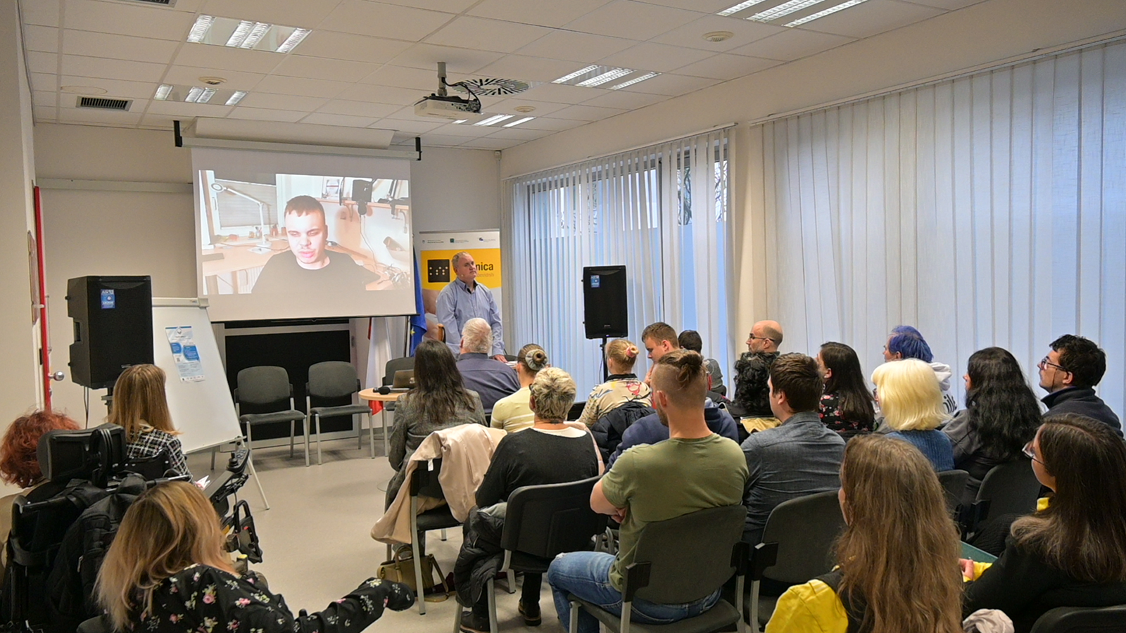 Audience watching a speaker on a screen during a hybrid event in a conference room.